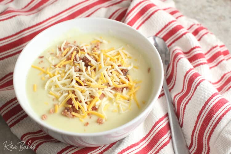 White bowl filled with cream cheese potato soup topped with shredded cheese and crumbled bacon. The bowl is sitting on top of a red and white striped towel with a silver spoon next to it.