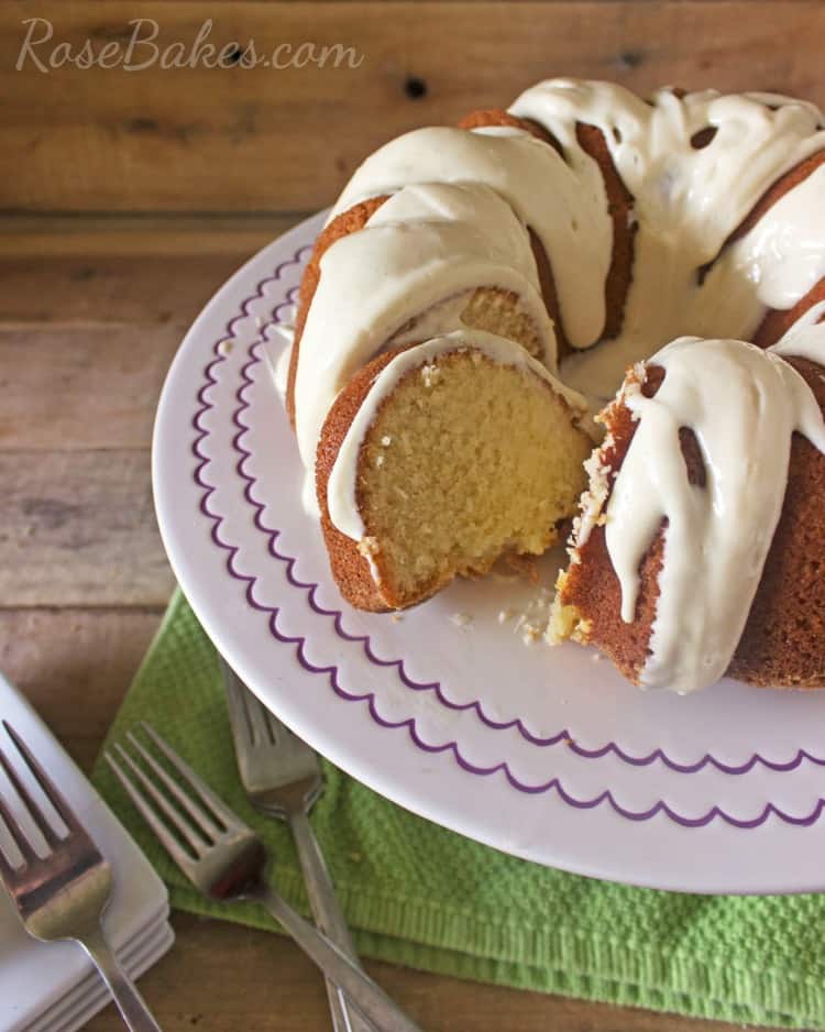 Sliced Pound Cake on a white cake stand with purple scallops. Below the cake stand is a green cloth napkin with forks.
