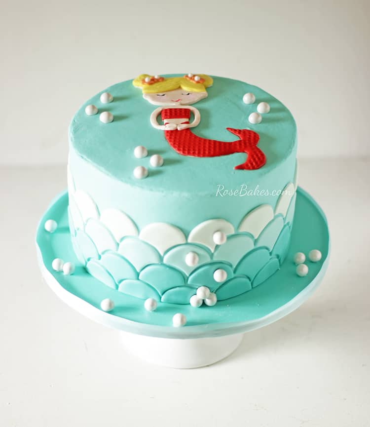 Sweet small mermaid cake. Teal cake with ombre scales up the sides and a 2d mermaid with a red tail on top. Pearls dot the top and sides.