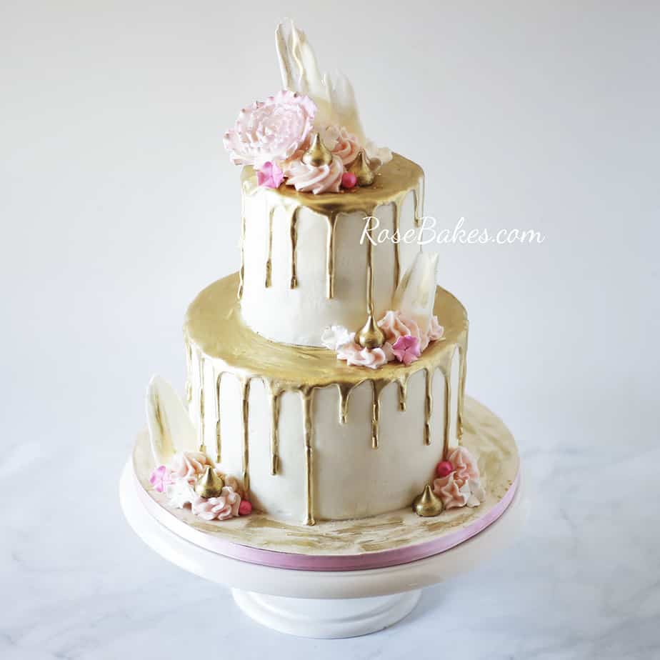 Two tiered white cake with gold drip on both tiers. On the tops and sides of each tier are pink flowers, white chocolate shards, and gold hersey kisses.