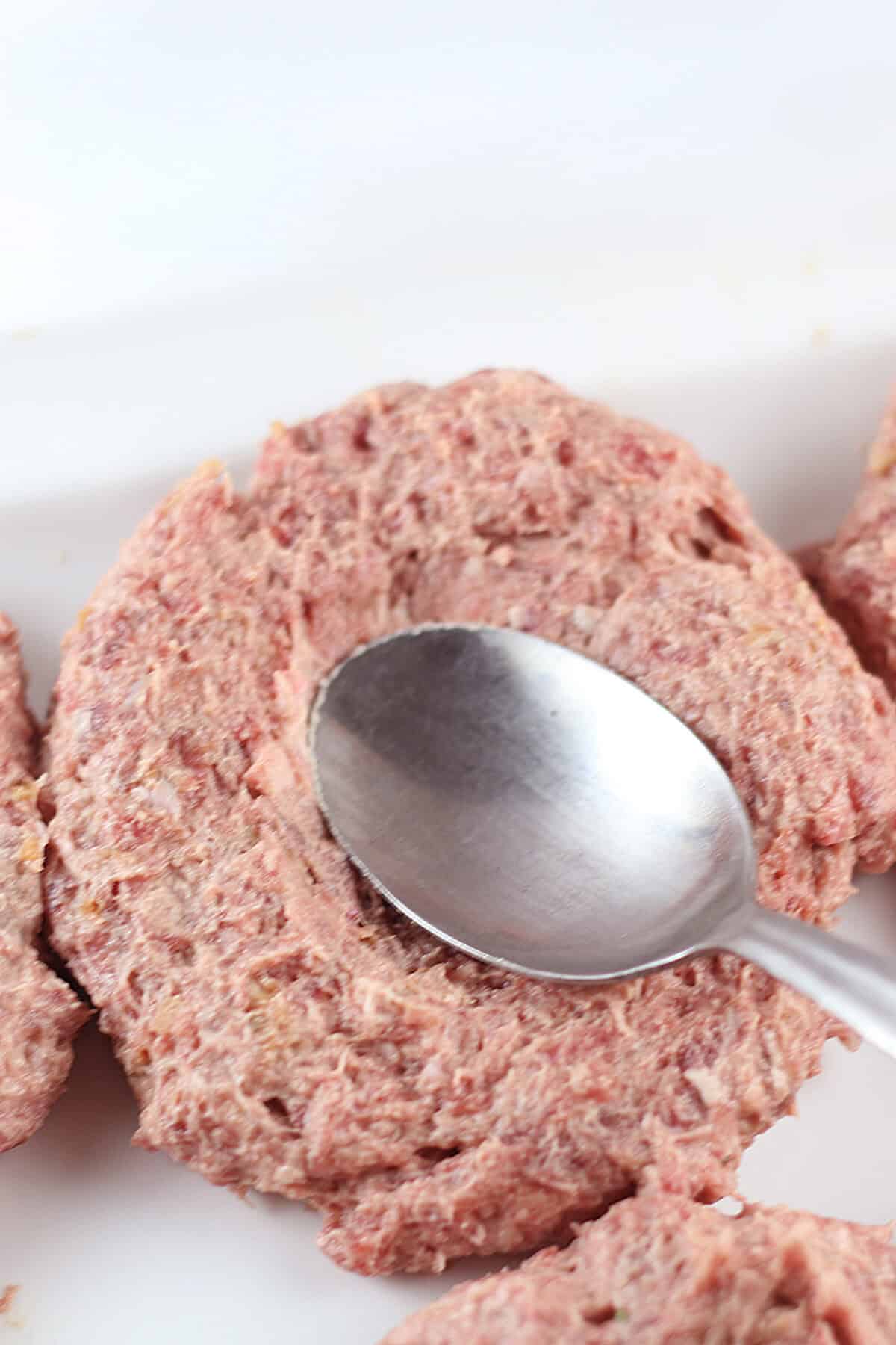 a spoon on a raw burger pressing an indention in the center