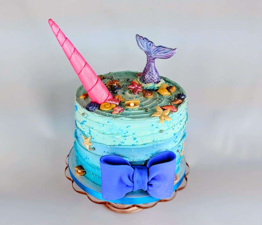 Ombre Blue Buttercream cake with a unicorn horn and a mermaid tail coming out of the top of the cake.
A blue bow is on the front.