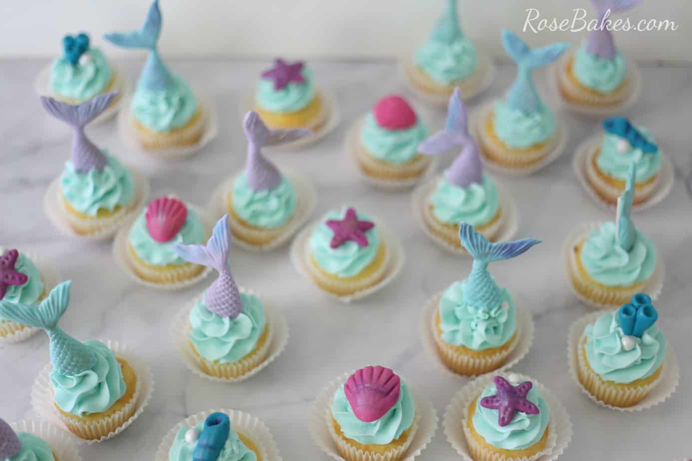 Vanilla cupcakes with light blue frosting. The cupcakes are topped with fondant mermaid tails and shells.