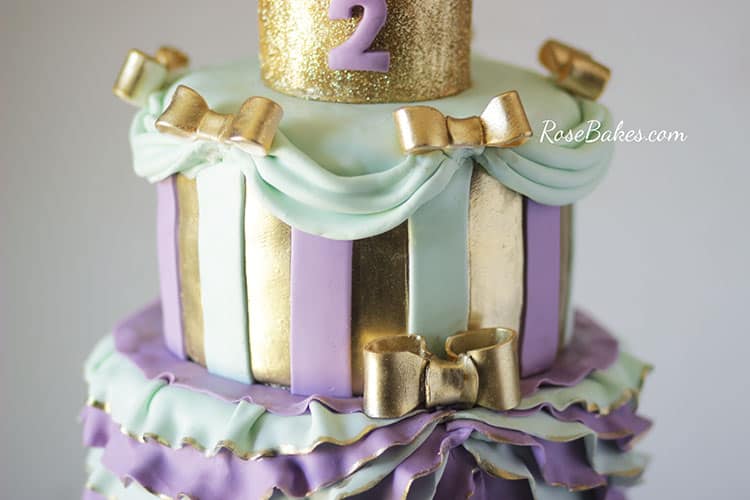 top tier of princess cake with stripes and gold bows and draping fondant