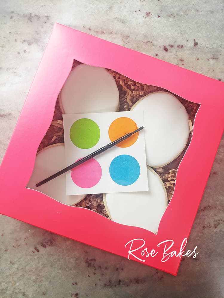 A pink box of blank cookies with a palette and paintbrush for Paint-Your-Own Cookie Sales..