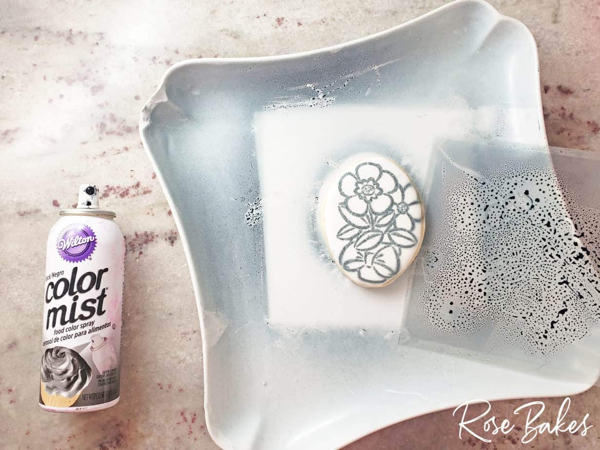 A stenciled PYO cookie with black color mist spray and a stencil on a white plate