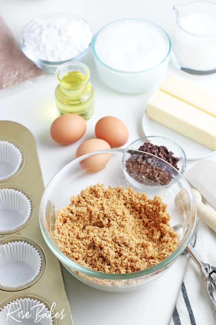 mixing bowl with graham cracker crumbs, melted butter and sugar plus other ingredients in background for s'mores cupcakes