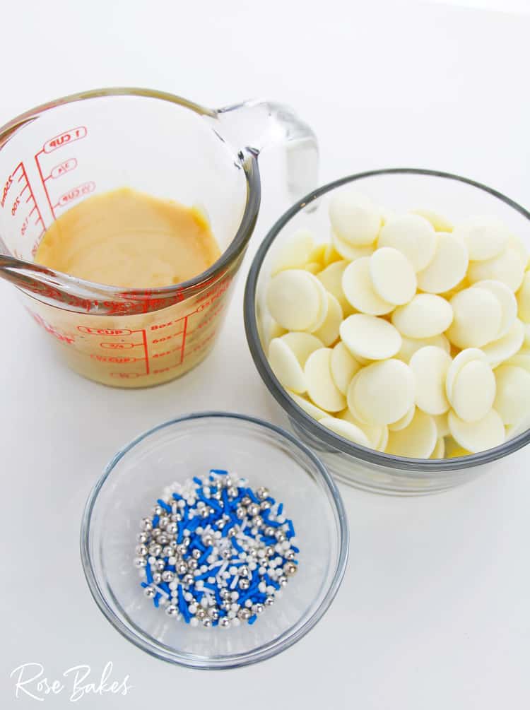 Sweetened condensed milk in a measuring cup, bowl of white chocolate wafer, and a small bowl of blue, white, and silver sprinkles