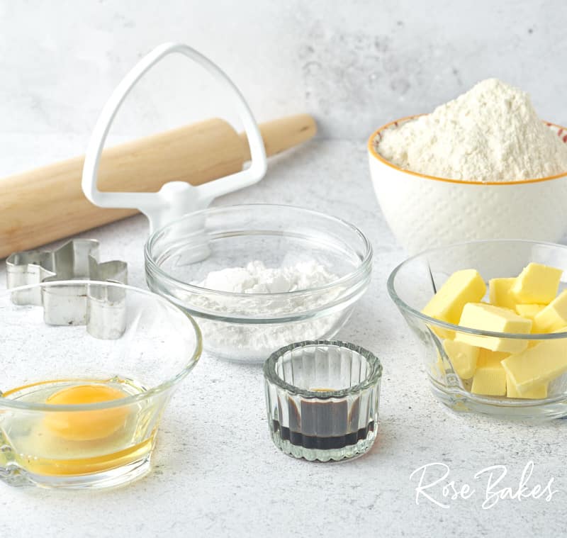Bowls of ingredients for the cookies measured out into white and glass mini bowls. Wooden rolling pin in background.