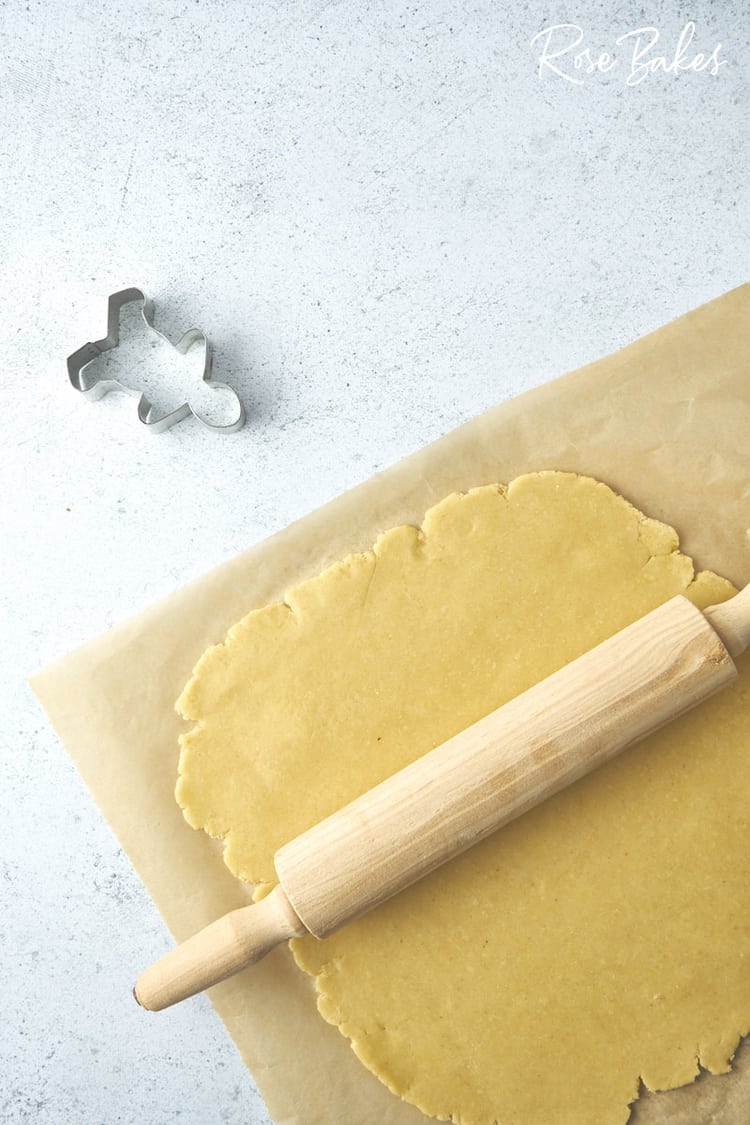 the cookie dough rolled out on a sheet of parchment. A rolling pin sits on top of the dough and a gingerbread man cookie cutter is off to the side.