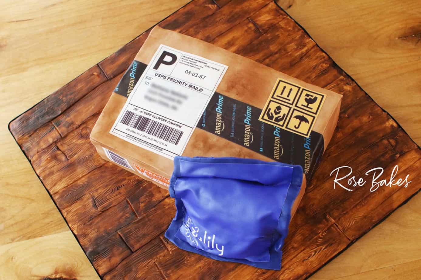 Higher up Back view of a cake shaped like and amazon cardboard shipping box and a blue zulily package. The cake is sitting on a cake board that looks like wood made from fondant and hand painting.