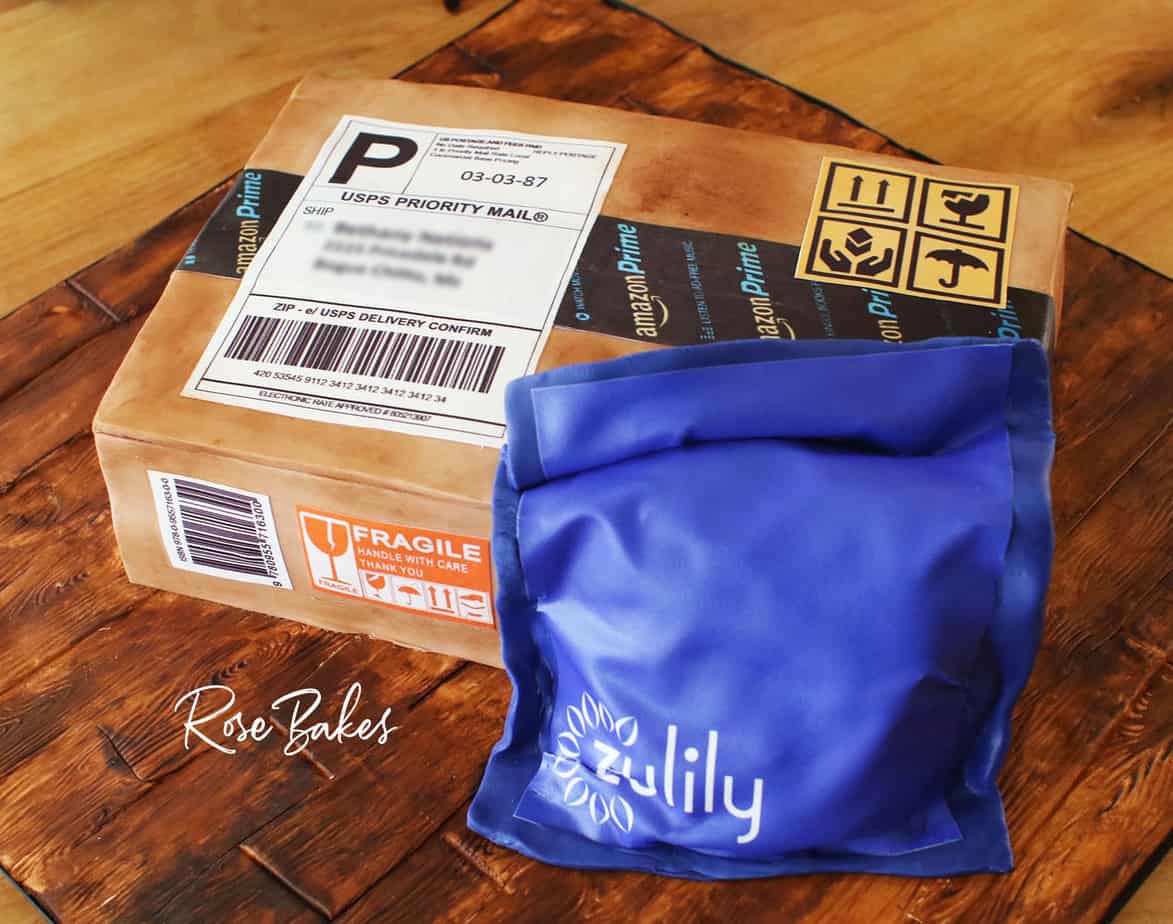 Cake shaped like and amazon cardboard shipping box and a blue zulily package. The cake is sitting on a cake board that looks like wood made from fondant and hand painting.