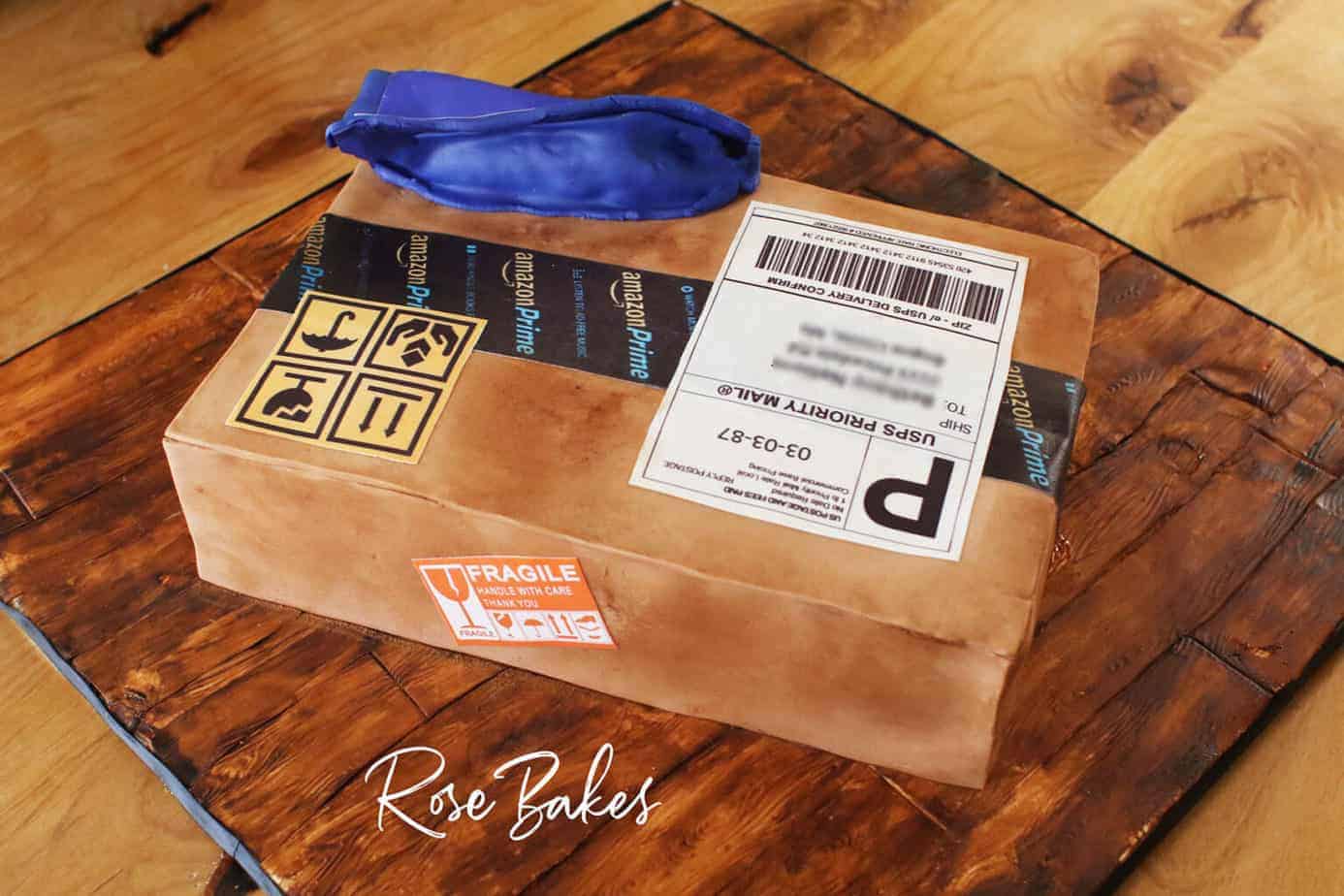 Back view of a cake shaped like and amazon cardboard shipping box and a blue zulily package. The cake is sitting on a cake board that looks like wood made from fondant and hand painting.