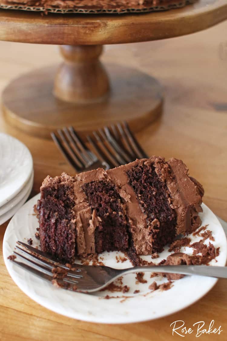 A slice of chocolate cake filled with chocolate frosting on a white plate. A fork is resting on the plate and a couple of bites have been eaten from the slice of cake. The wooden cake stand is in the background.