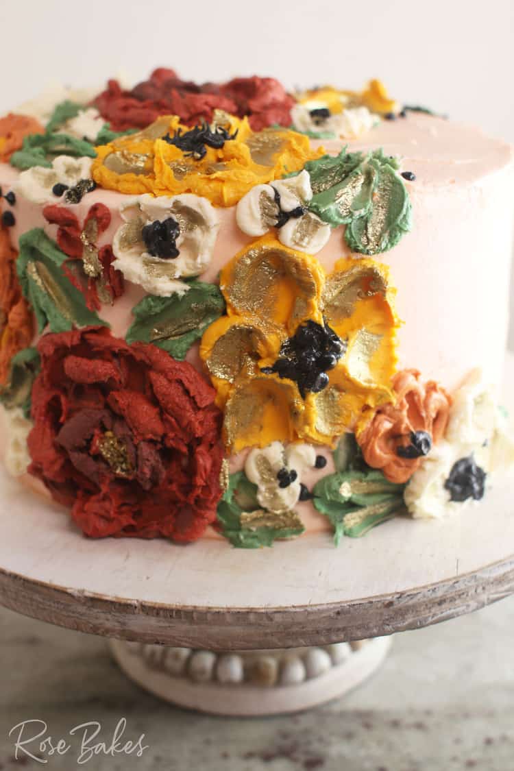 Close up view a cake frosted white with green leaves and red, yellow, and white buttercream flowers created with palette knives. The cake is displayed on a rustic wood cake stand.