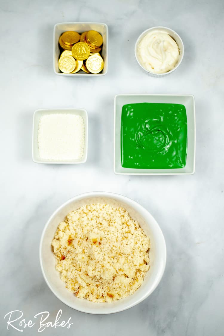 bowls of everything needed to make the St. Patrick's Day cake pops. Top left square bowl has gold coins, top right round bowl has frosting, middle left square bowl has white nonpareil sprinkles, middle right square bowl has melted green candy melts, bottom large round bowl has cake crumbs.