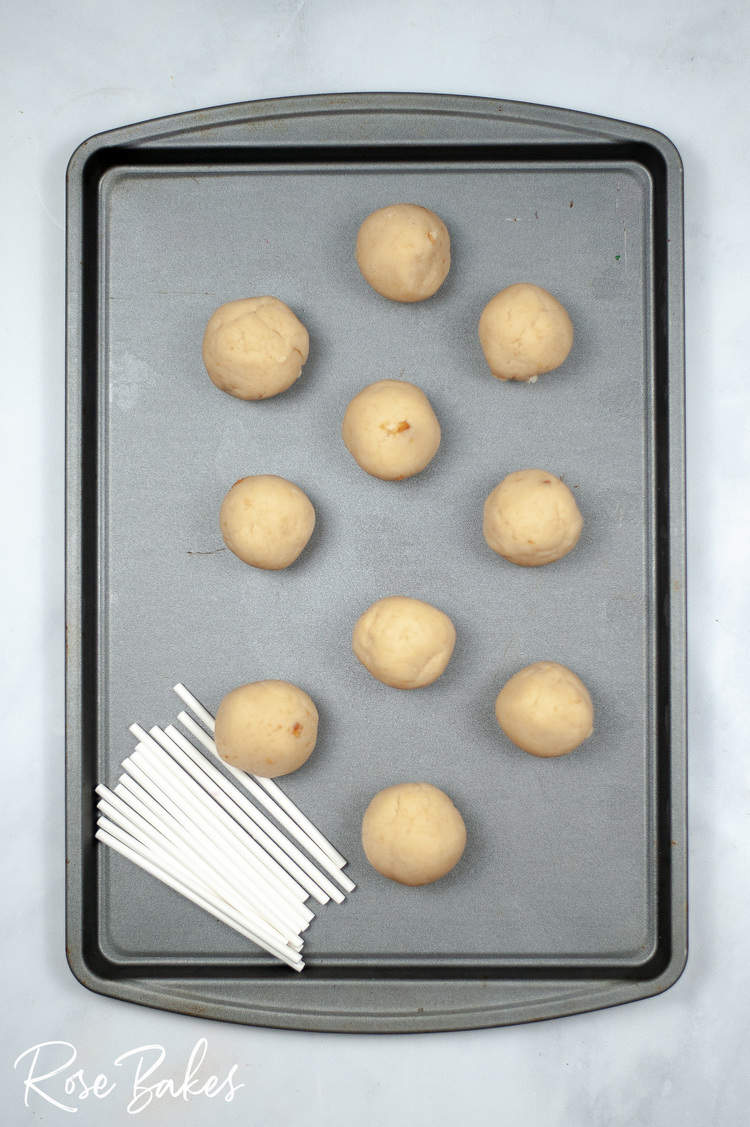 Cake pops rolled into balls on a baking sheet. White pop sticks are in a stack at the bottom of the pan.