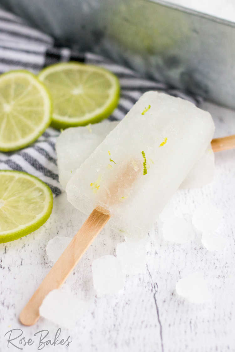 2 popsicles with lime zest displayed on crushed ice. Lime slices in the background.