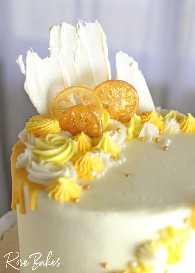 Close up of the yellow drip and yellow buttercream rosettes and stars on the top of the cake.  Among the buttercream decorations are candied lemons, white chocolate fans, and gold and white sugar pearls. 