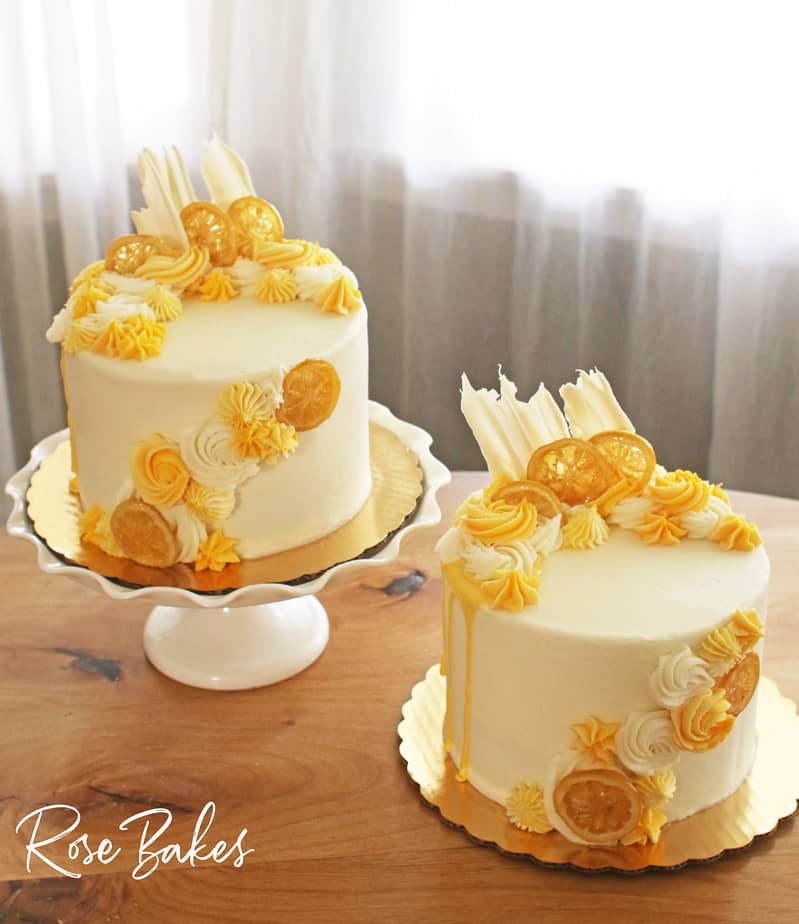 Two cakes frosted in white buttercream with yellow drip and yellow buttercream rosettes and stars around the top left and front right of the cake.  Among the buttercream decorations are candied lemons and white chocolate fans.  The cake on the left is displayed on a white cake stand with a large ruffled edge and the other is sitting on the wooden table.