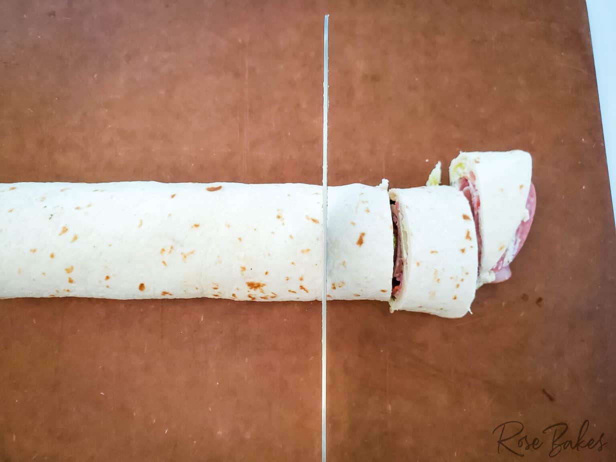 slicing italian pinwheels from rolled up tortilla that is stuffed with meats and cheese