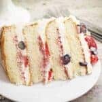 Slice of Chantilly Cake with berries in the filling and on top of the cake. The cake is on a round white plate.