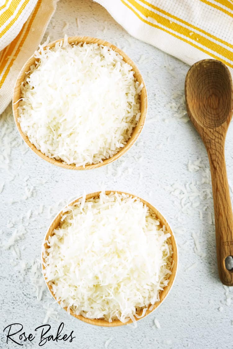 Two wooden bowls on the left filled with shredded coconut. Wooden spoon is to the right of the bowls