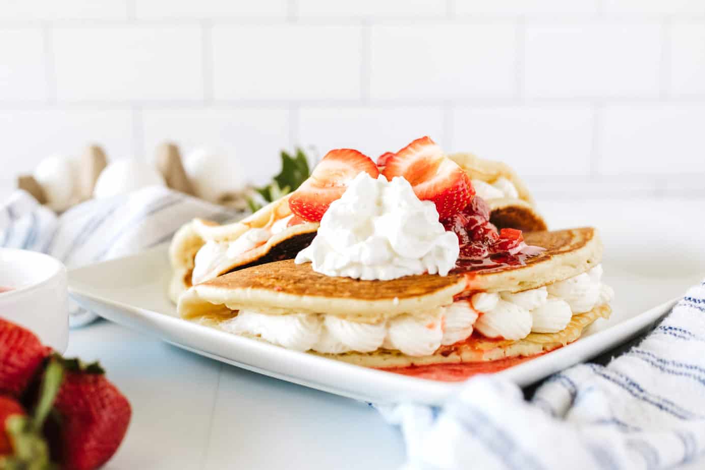 two buttermilk pancakes stuffed with no-bake cheesecake filling folded like tacos on a white plate with fresh strawberries and strawberry syrup on top