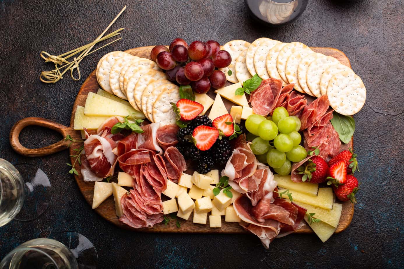 Charcuterie board filled with meats, cheeses and fruits with wine glasses next to it on a black counter