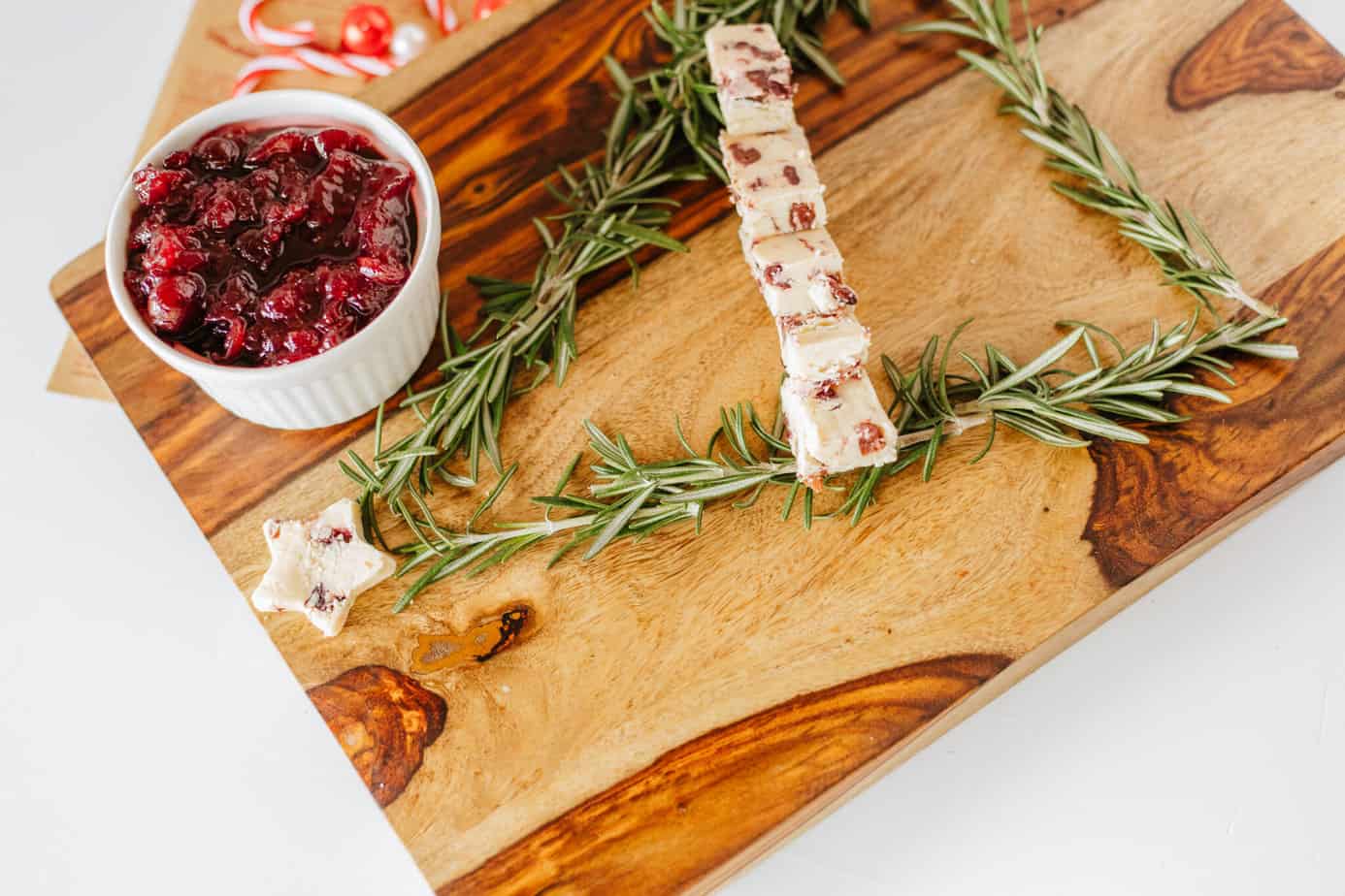 cutting board with rosemary sprigs and sliced cheese in shape of christmas tree on a cutting board with bowl of cranberry sauce
