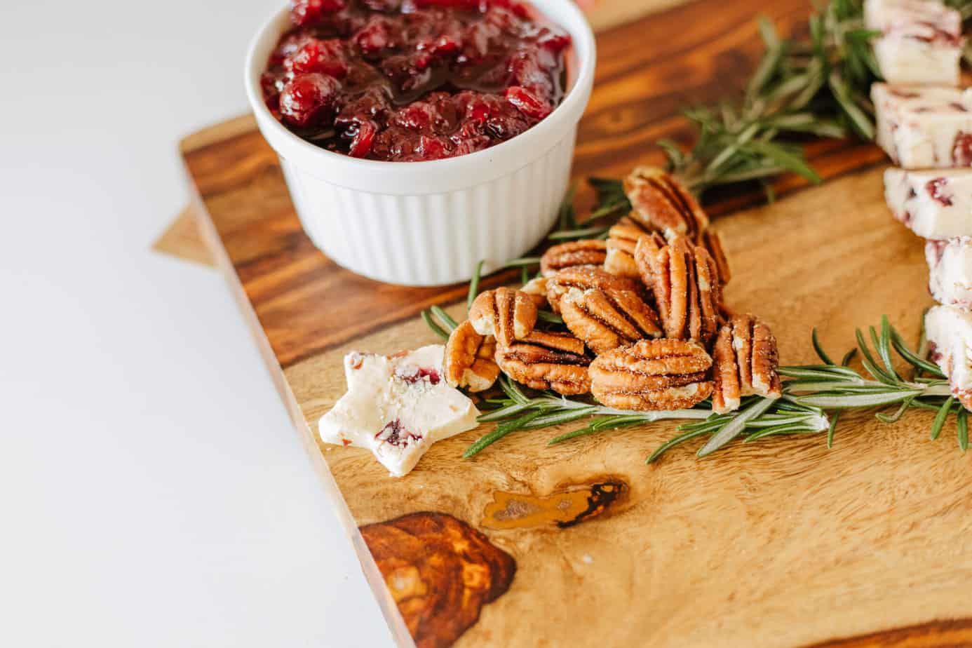 adding pecans to cutting board with rosemary sprigs in shape of christmas tree on a cutting board with bowl of cranberry sauce