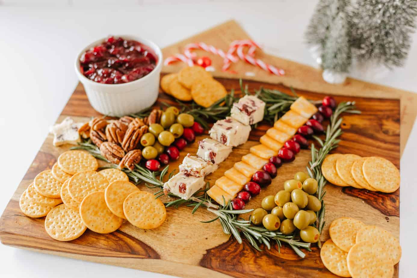 adding olives to cutting board with rosemary sprigs in shape of christmas tree on a cutting board with bowl of cranberry sauce