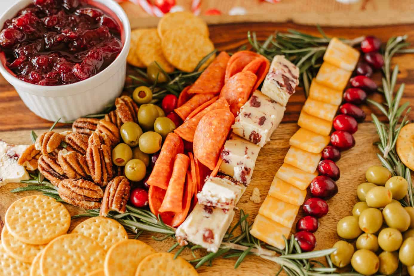 adding pepperoni to cutting board with rosemary sprigs in shape of christmas tree on a cutting board with bowl of cranberry sauce for Christmas Charcuterie board