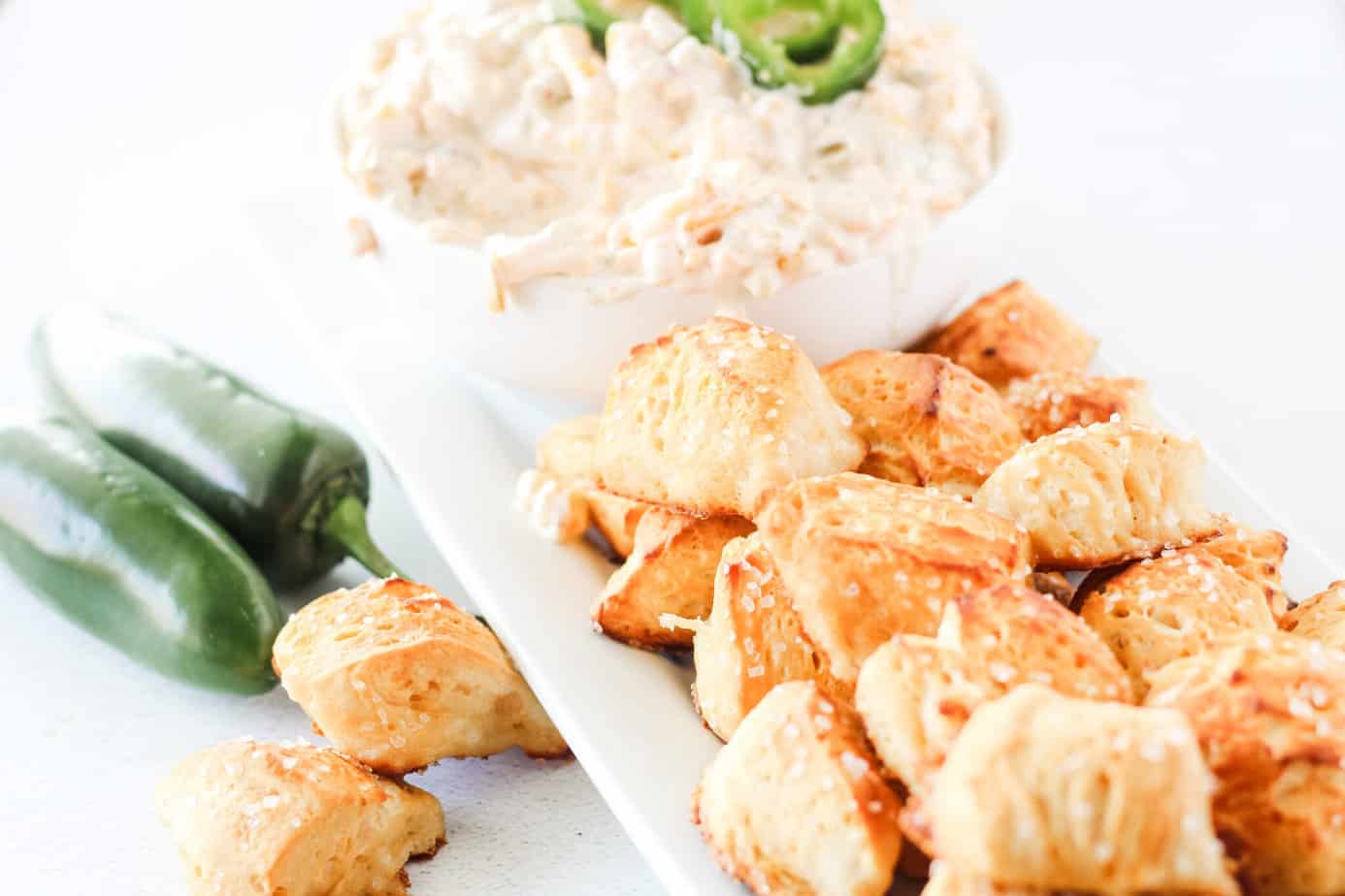 Biscuit Pretzel Bites on a white platter with jalapeno peppers on the side and a dip in the background