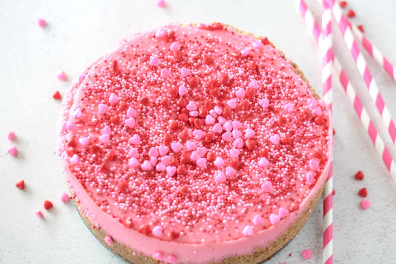 hot pink valentine's day cheesecake with red pink and white sprinkles on a white surface with pink striped straws