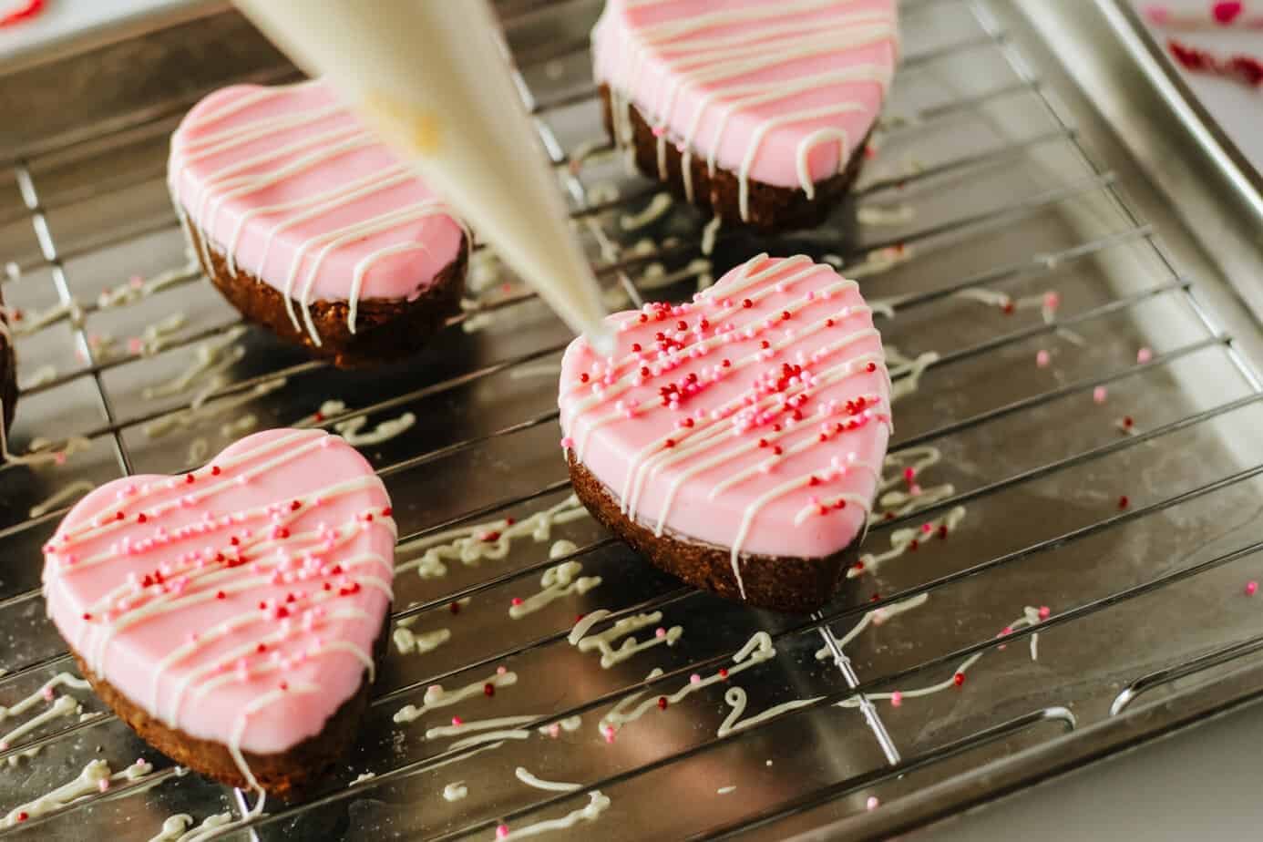 drizzling white chocolate and sprinkles over Heart Brownies on a wire rack with pink chocolate on top