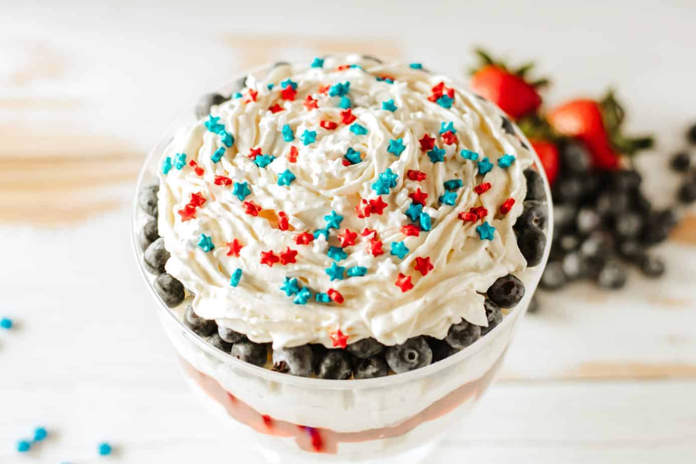 layers of pudding mixture with angel food cake and strawberries in trifle dish with blueberries on top and sprinkles