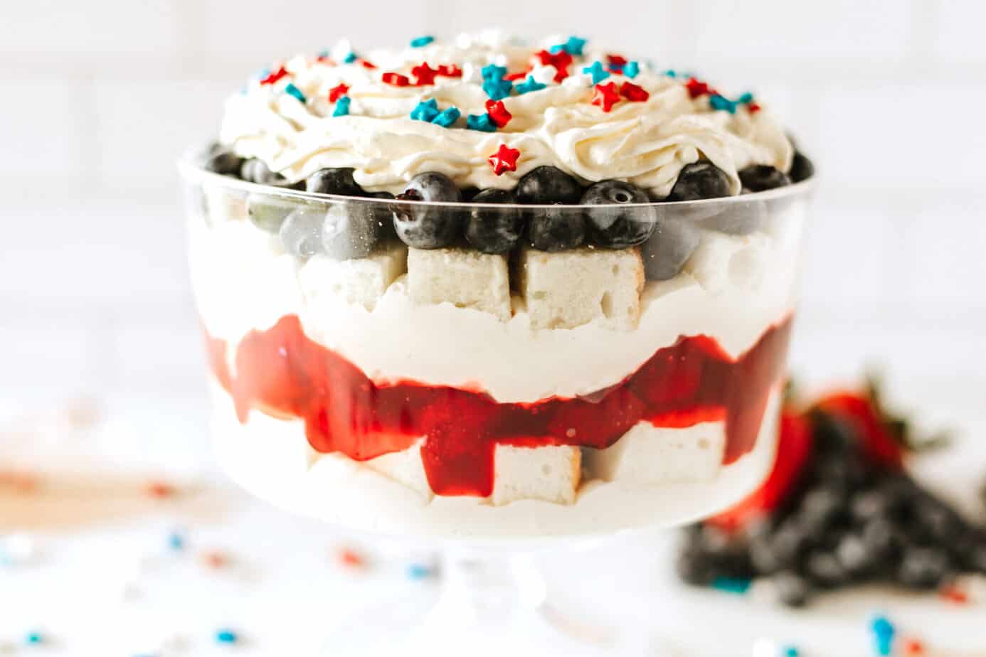 Red, White and Blue Trifle in a trifle dish with star sprinkles on top and berries in the background
