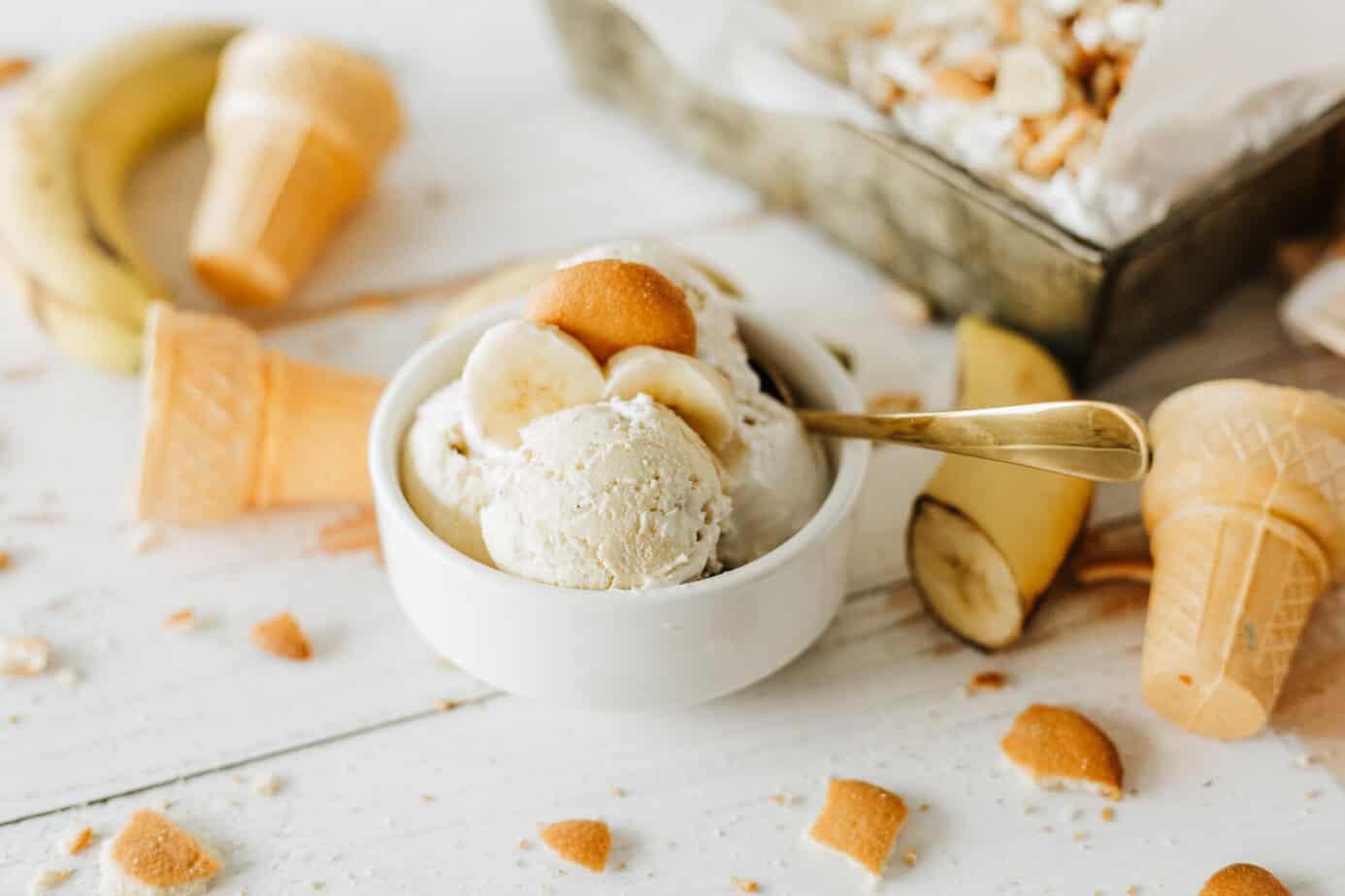 Banana Pudding Ice Cream on a white surface with broken Nilla Wafers, sugar cones and cut bananas