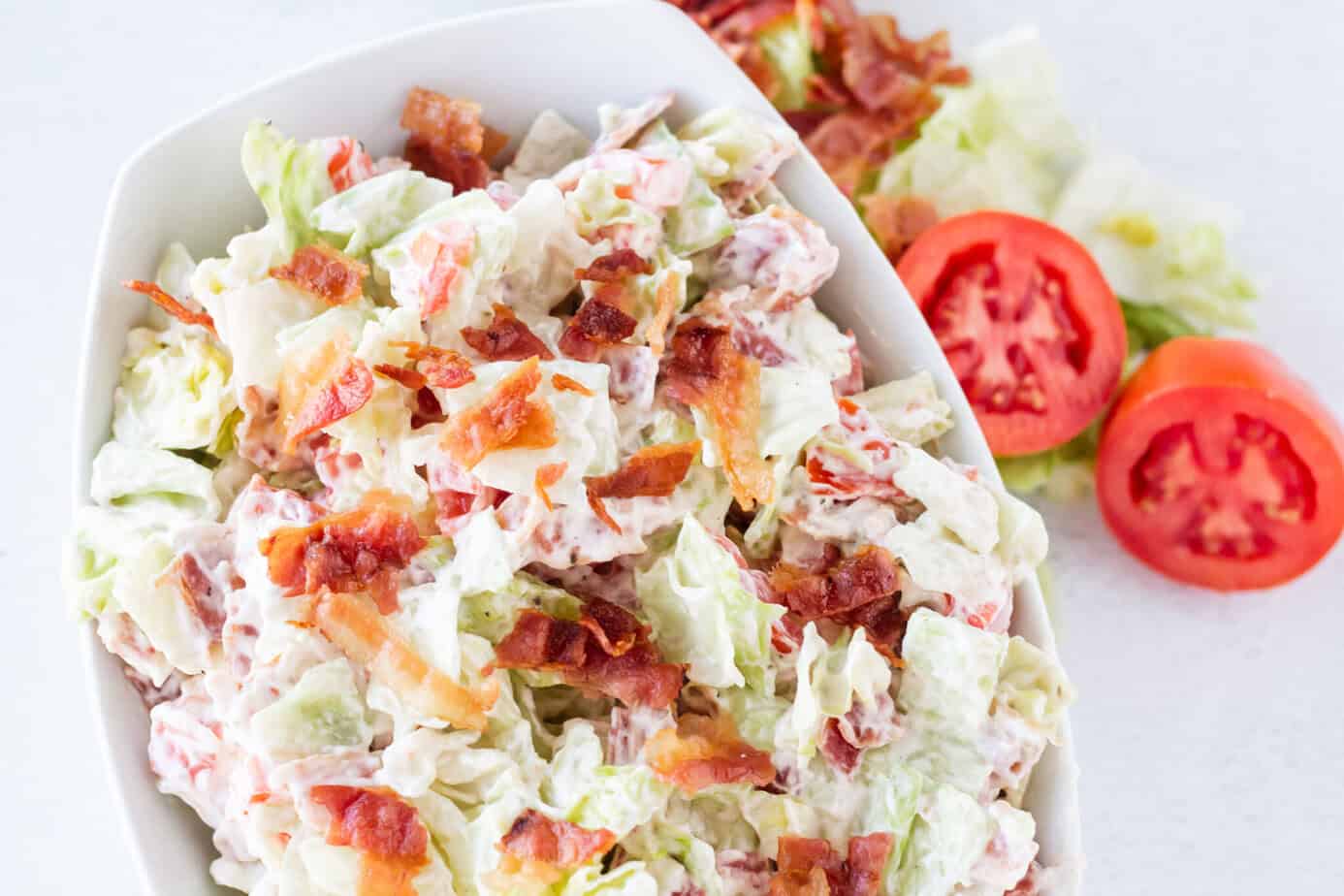 BLT Salad in white bowl on white surface with bacon lettuce tomatoes on the table next to the bowl