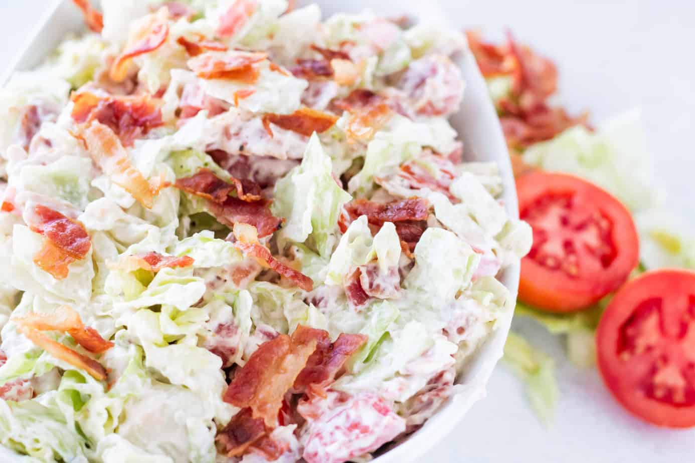 BLT Salad in white bowl on white surface with bacon lettuce tomatoes on the table next to the bowl