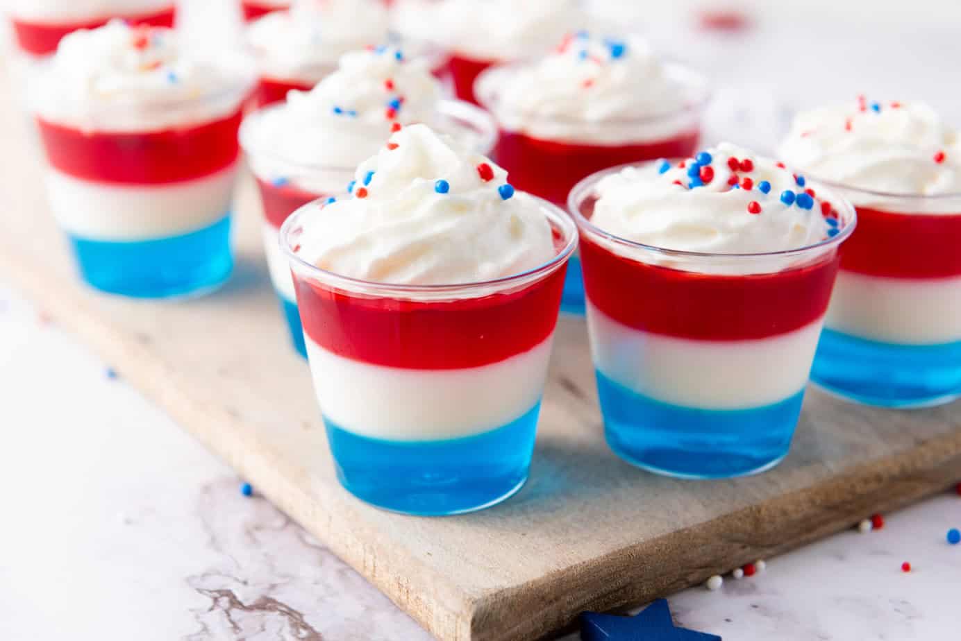 red white and blue layered jello shots whipped cream and sprinkles on top sitting on a wooden cutting board