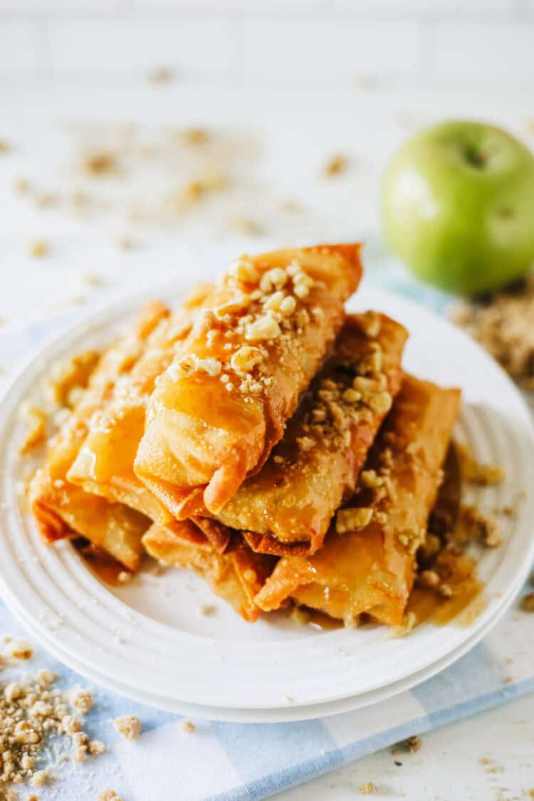 apple pie egg rolls stacked on a plate with caramel and struesel and walnuts