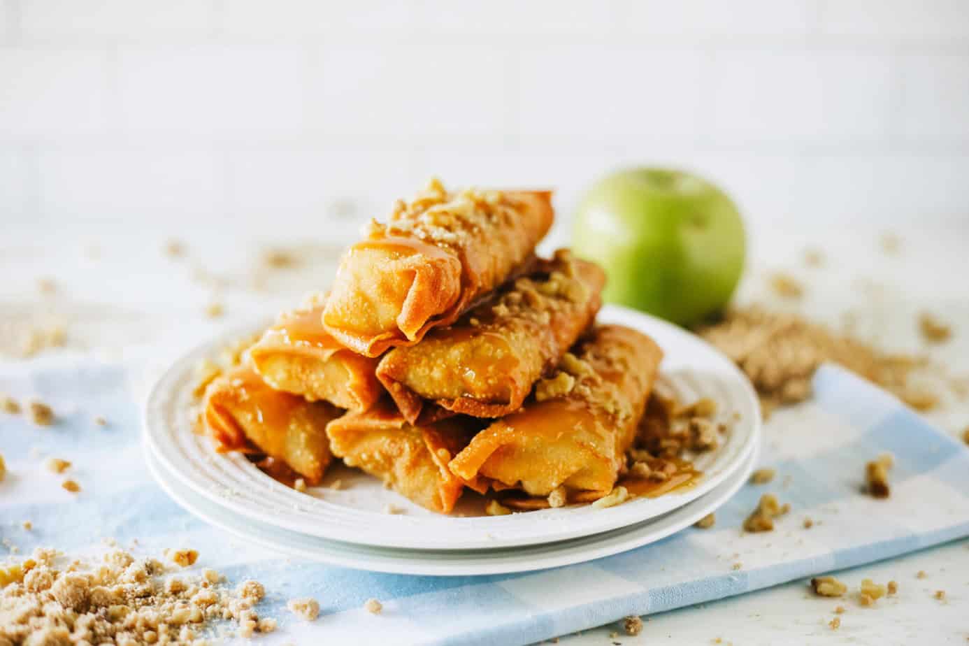 apple pie egg rolls stacked on plates with caramel, walnuts and streusel 