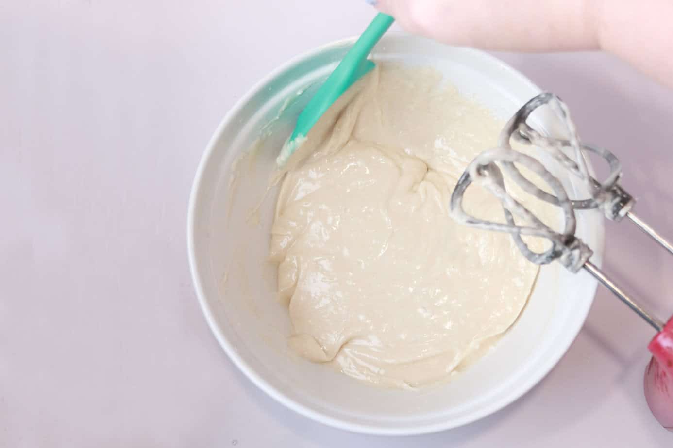 scraping down the sides of a bowl with cake goop in it and a mixer sitting beside it