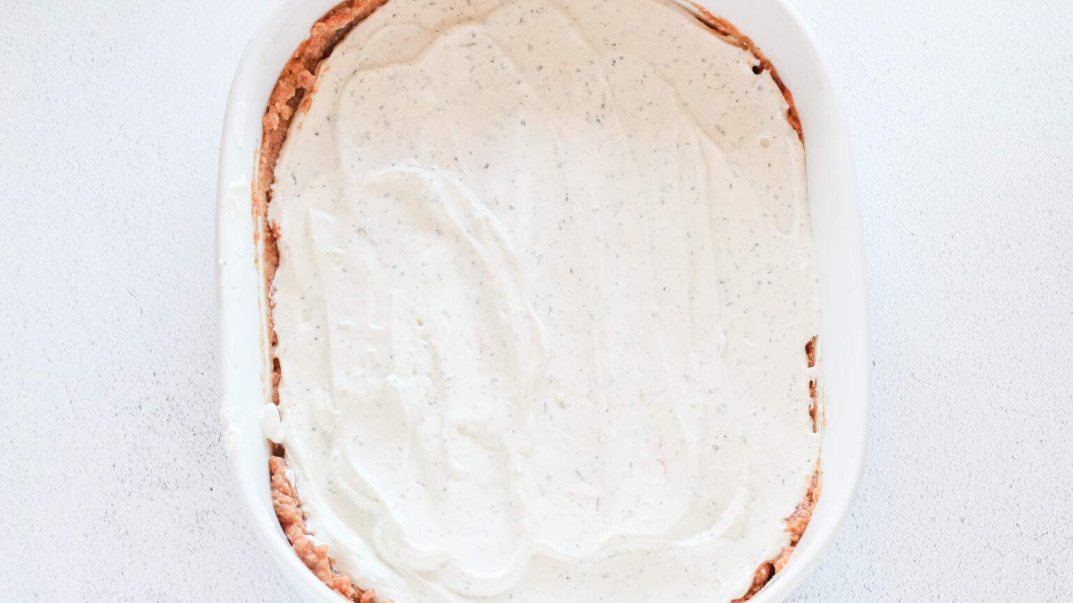 white oval baking dish with beans and salsa mix spread in bottom and sour cream mixture spread over top