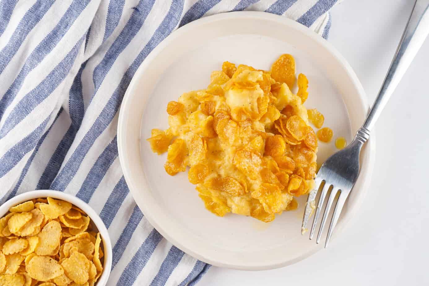 cooked Funeral Potatoes on a white plate next to a bowl of cornflakes
