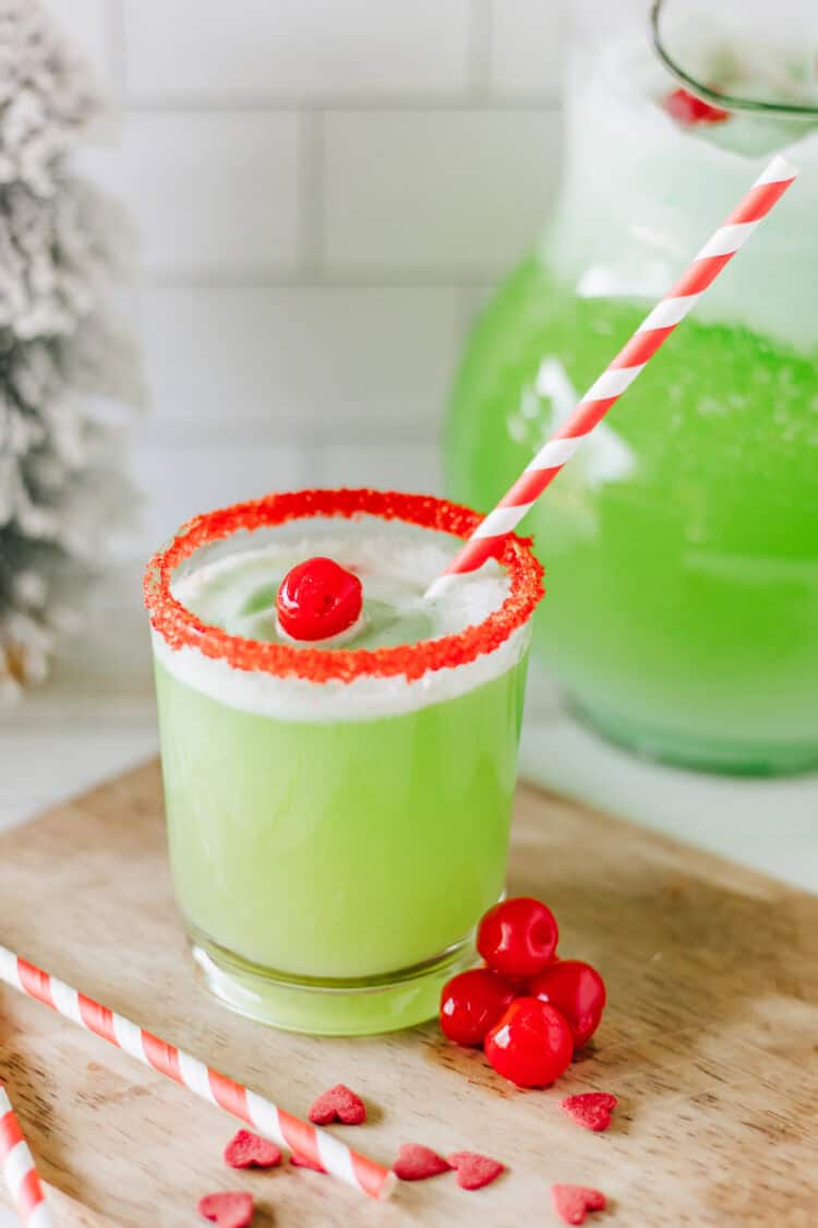Grinch punch in a small glad with red sugar on the rim of the glass and a striped straw and some maraschino cherries