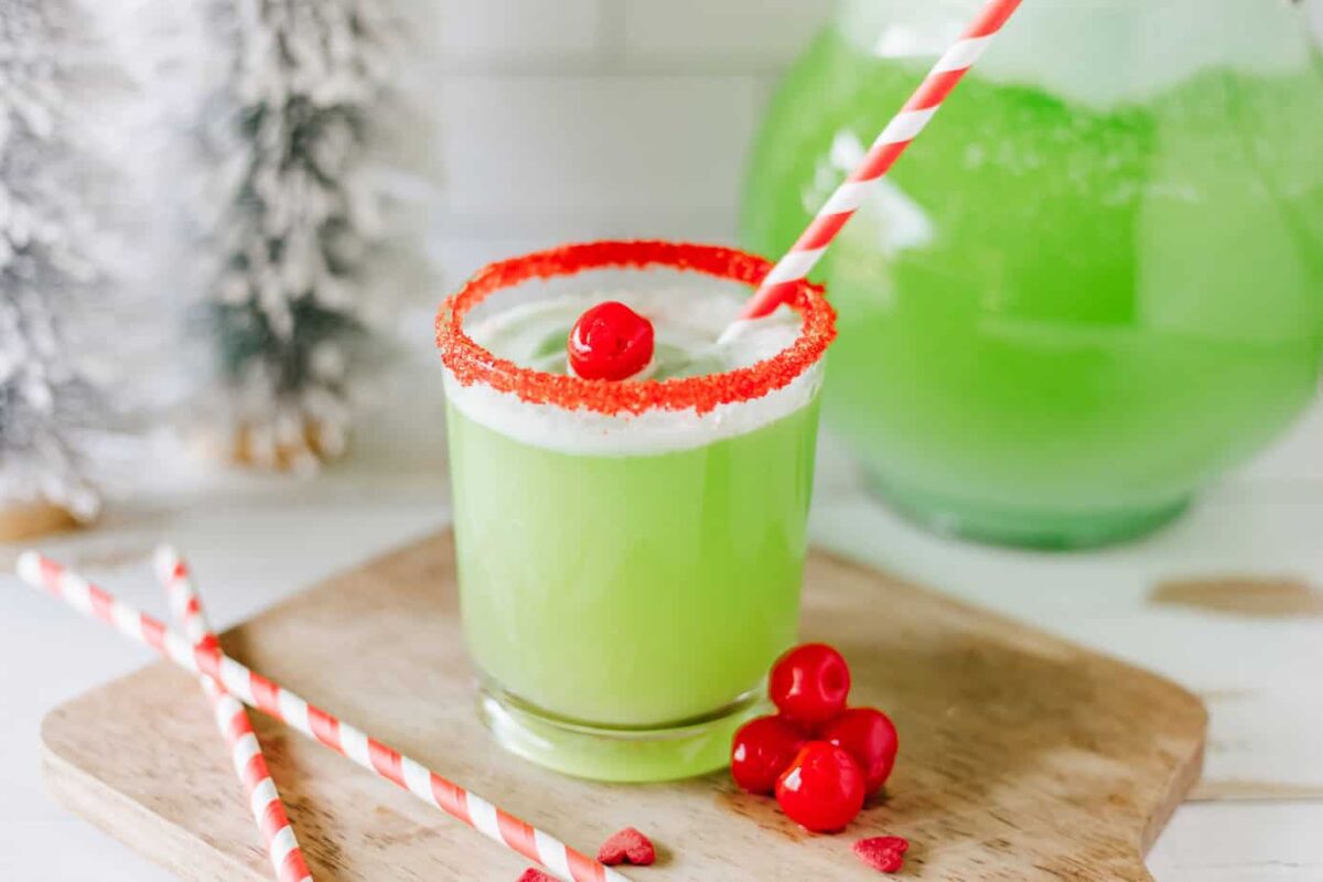 Grinch punch in a small glad with red sugar on the rim of the glass and a striped straw and some maraschino cherries