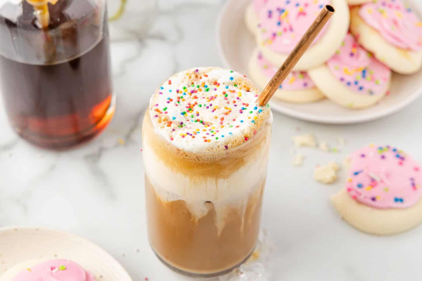 Sugar Cookie Latte with colorful sprinkles on top. A plate of pink frosted sugar cookies is to the right and a jar of the sugar cookie syrup is to the left in the background